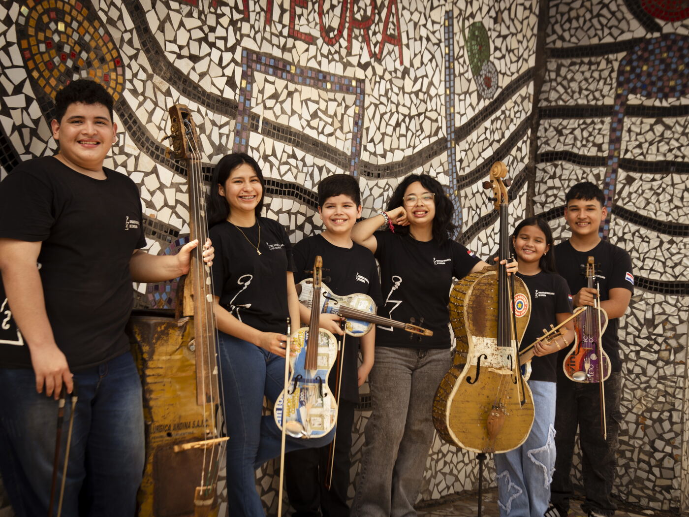 Die Protagonistinnen und Protagonisten lachen mit ihren Instrumenten vor dem Mosaik der Orquesta de Reciclados de Cateura in die Kamera